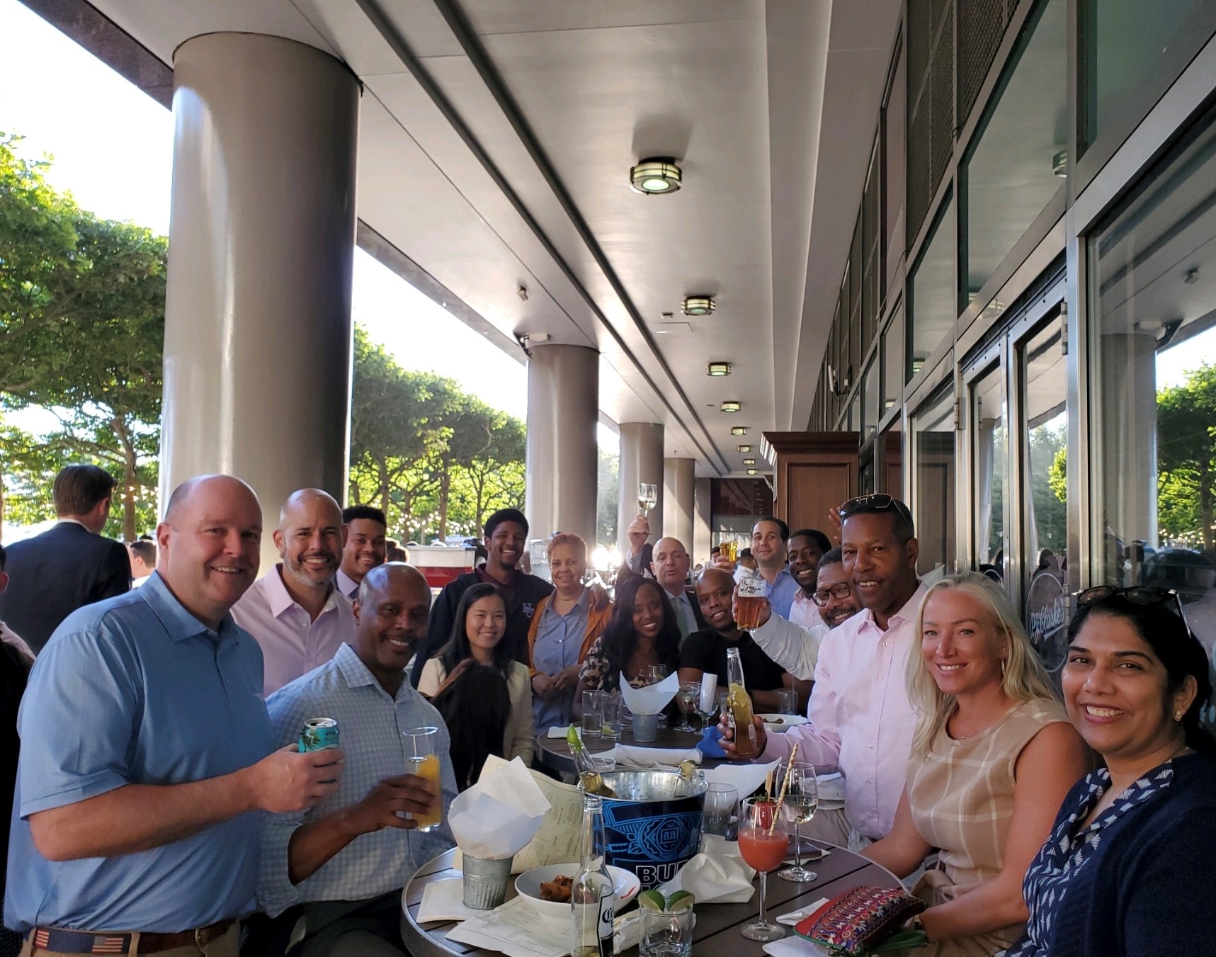 Group of diverse people smiling and holding drinks while seated around outdoor tables at a social gathering under a building overhang.