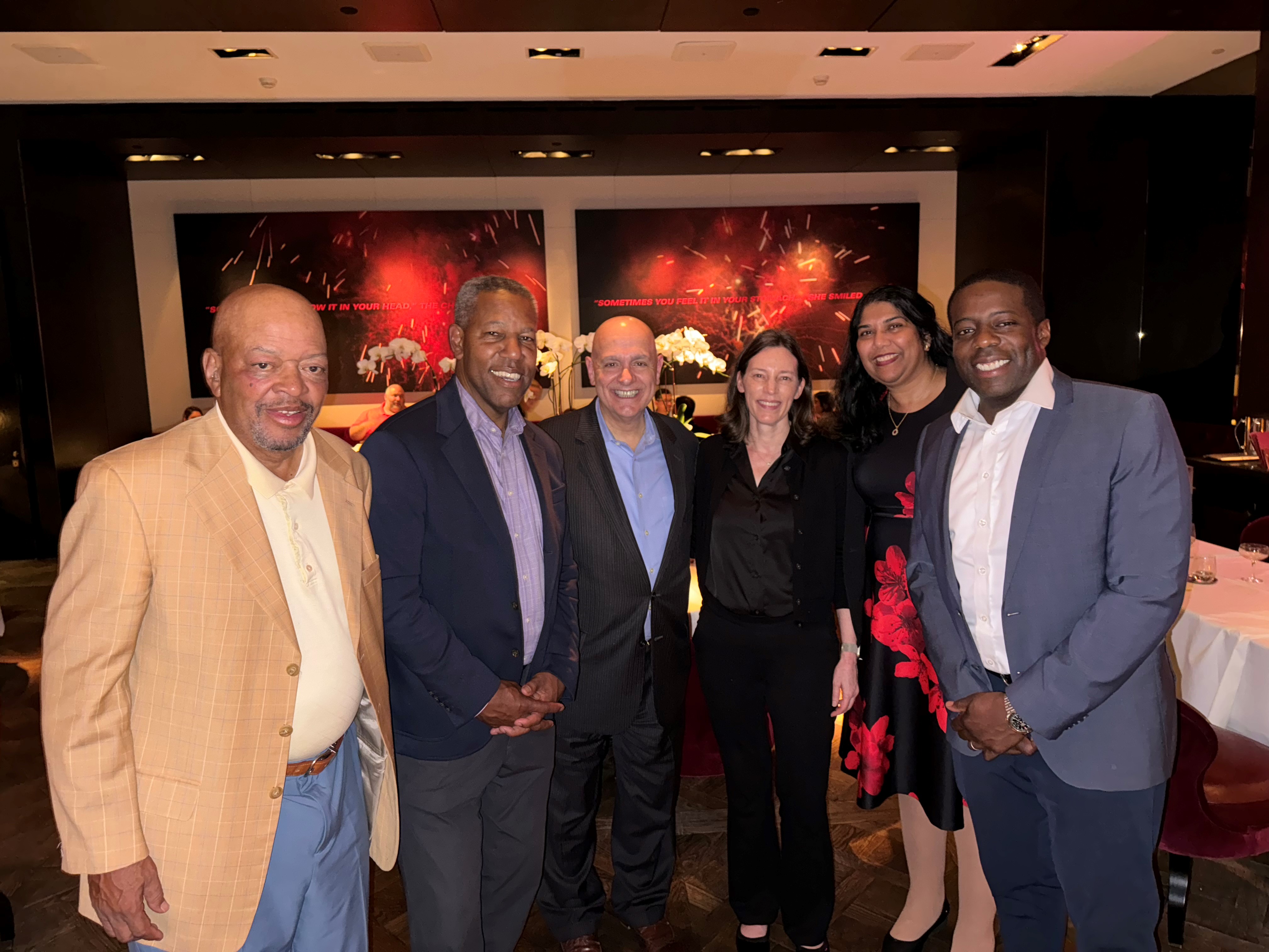 Six people standing and smiling together in a restaurant with dark walls and red artwork in the background.