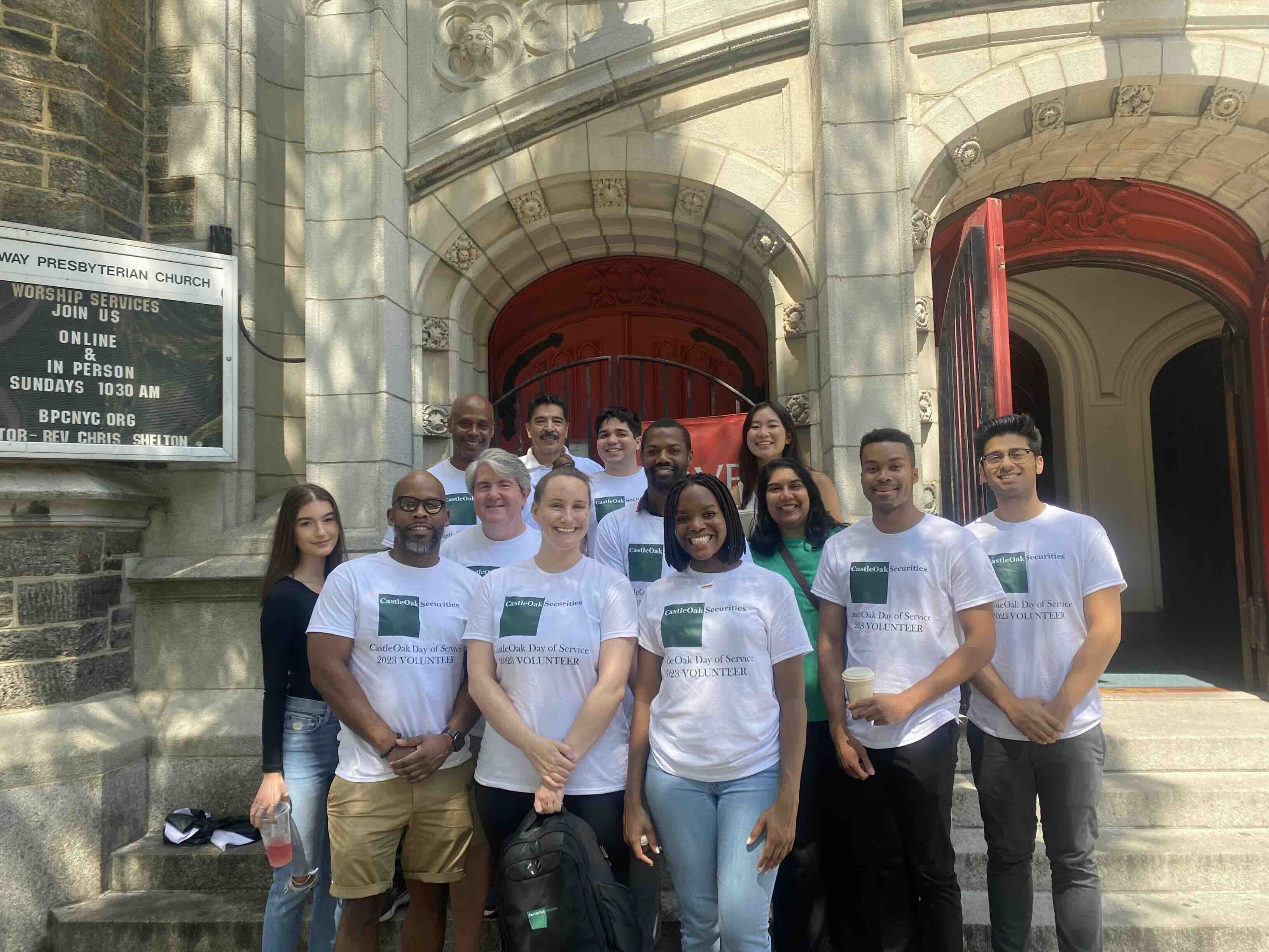 Group of diverse volunteers wearing CastleOak Securities Day of Service 2023 T-shirts standing and smiling outside a stone church with red doors.