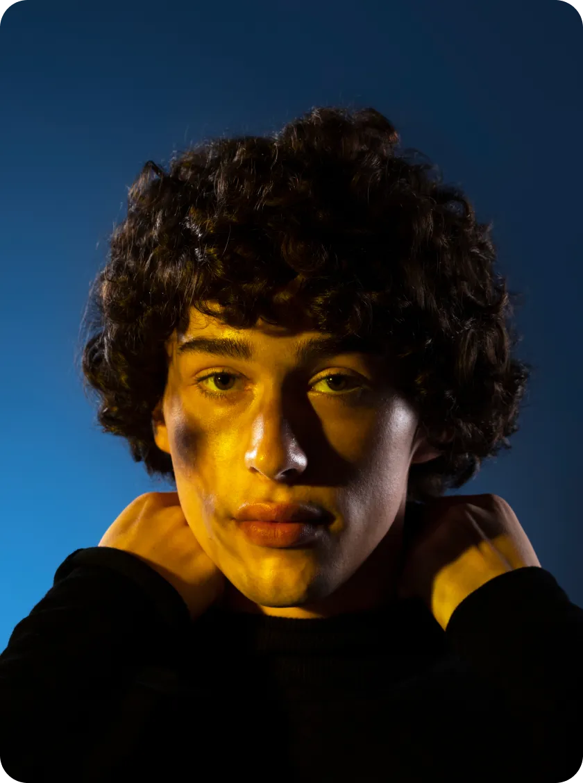 Close-up portrait of a person with curly hair and yellow light casting shadows on their face against a dark blue background.