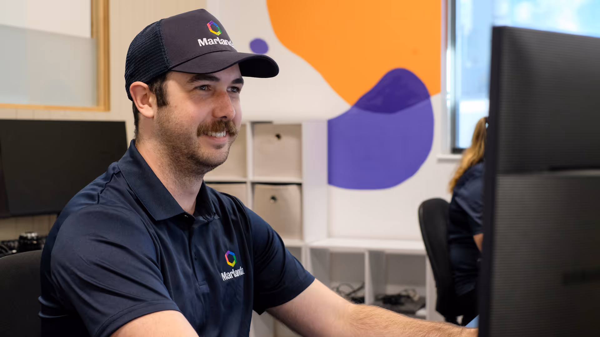 Man wearing a Marlanda cap and shirt sitting at a desk working on a computer in a colourful office.