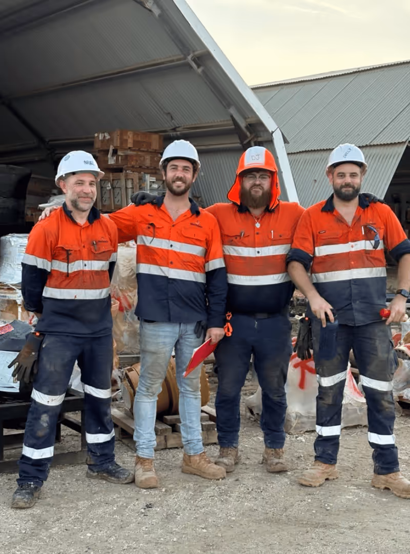 Smiling worker in an orange and navy Marlanda safety uniform and white hard hat standing behind a large empty plastic container inside a storage warehouse.