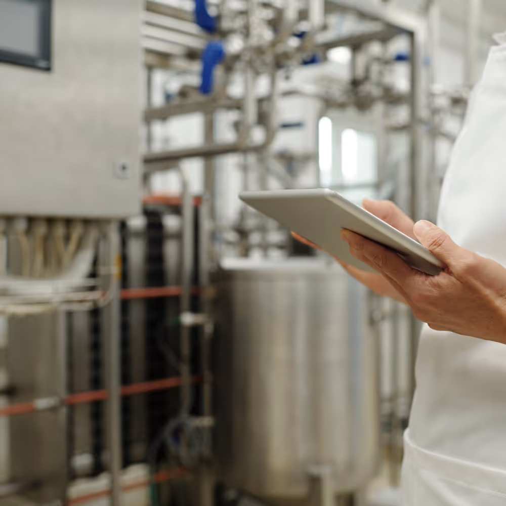 Person holding a tablet in a food processing facility with stainless steel equipment and pipes in the background.