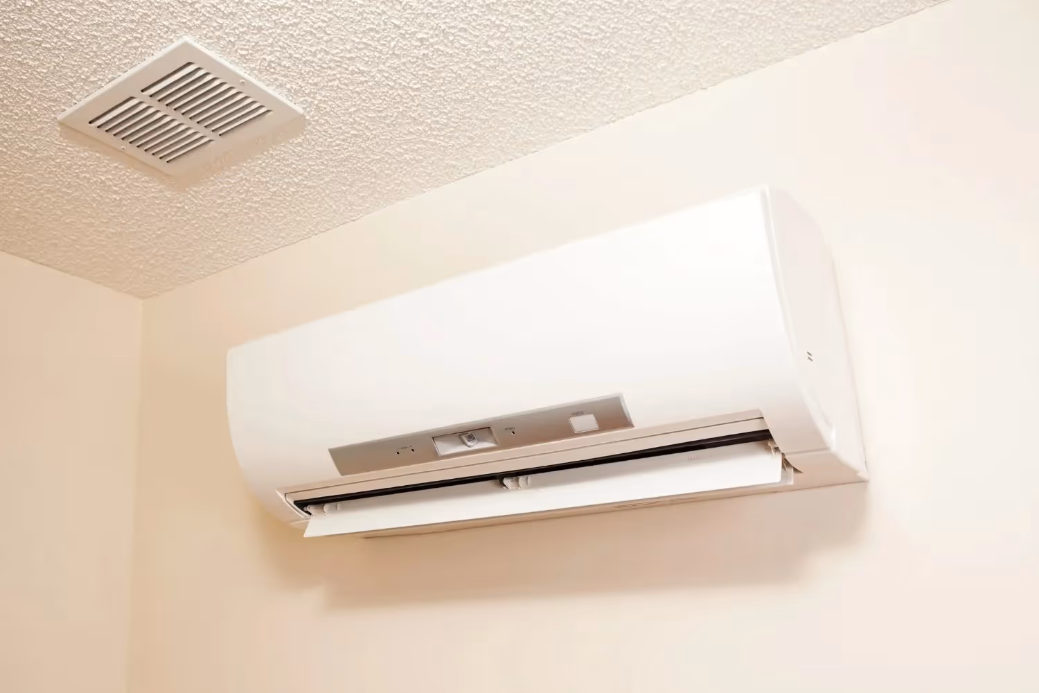 White ductless AC unit mounted high on a beige wall near a textured ceiling with a vent.