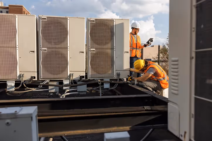 Two technicians in hard hats and safety vests are working on multiple commercial AC units on a rooftop.