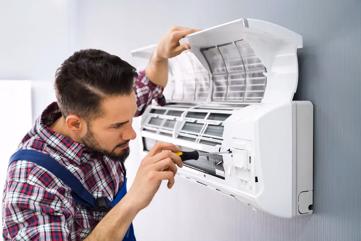 A technician with a beard and plaid shirt is using a screwdriver to fix a wall-mounted air conditioner.