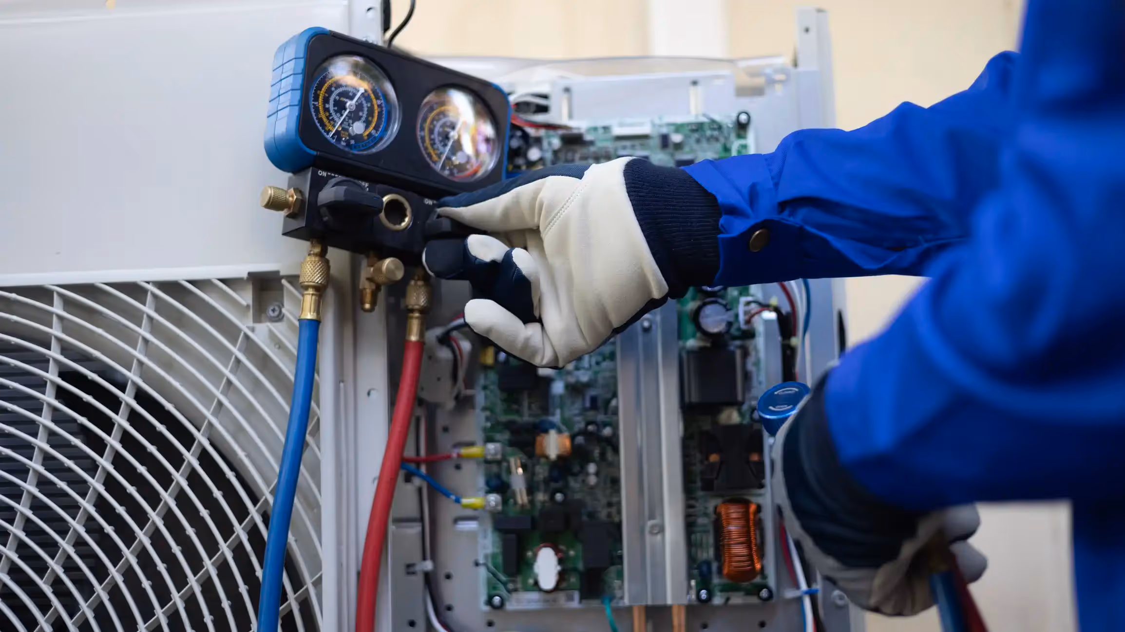  A close-up image shows a technician, wearing white and blue gloves, checking the pressure gauges of an air conditioner unit. The technician's hand is adjusting a valve on the manifold gauge set, which is connected by blue and red hoses to the internal components of the AC unit.