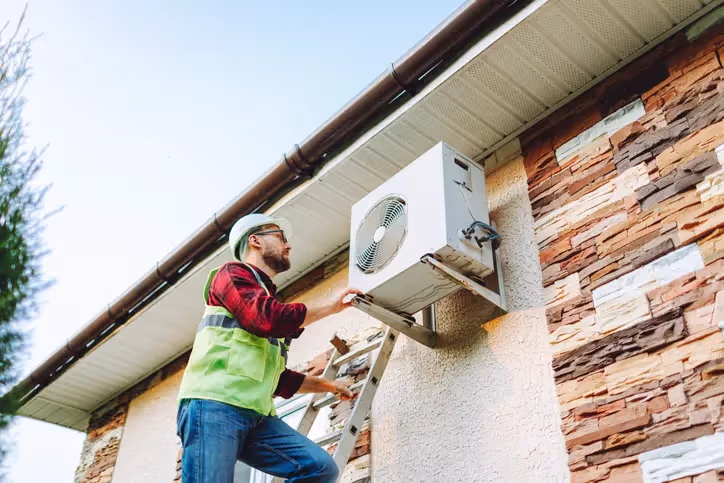 HVAC technician on a ladder installing a heat pump on the side of a house.