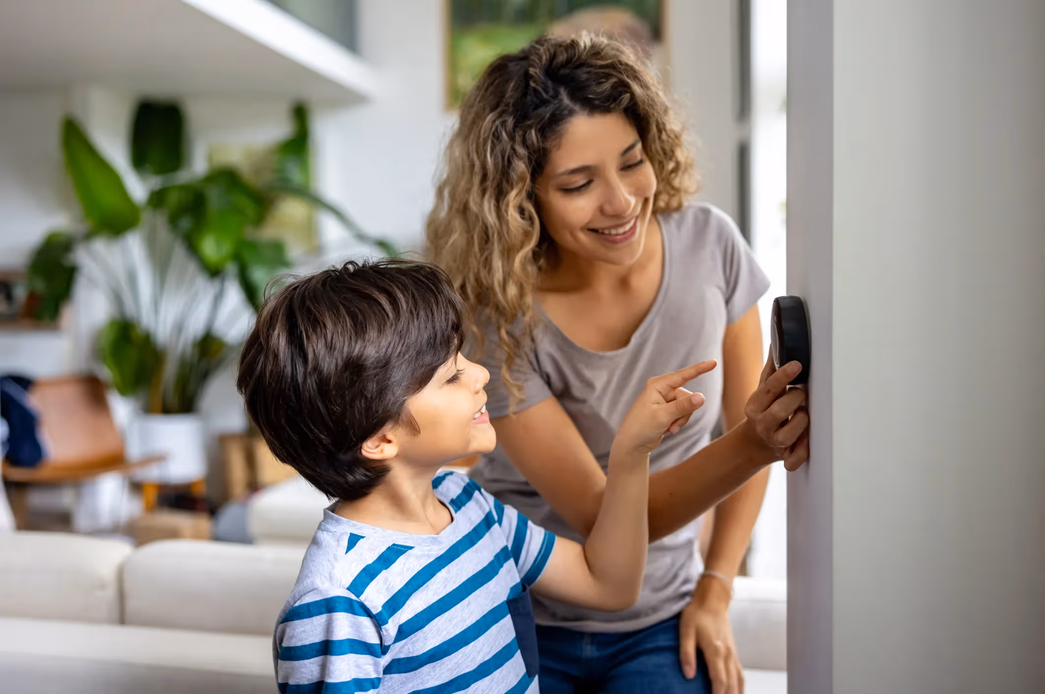 Smiling mother and son adjusting a round, modern smart thermostat on a living room wall.