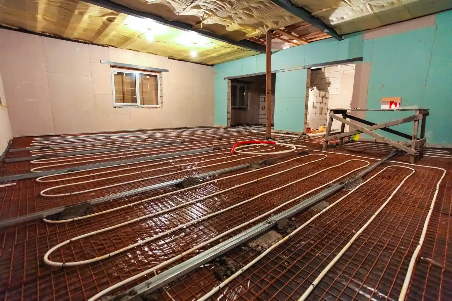 A room under construction has a network of white PEX tubing laid out on the floor for radiant heating.