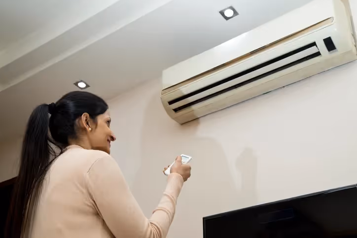 A person reaching up towards a wall-mounted mini-split air conditioner, holding a remote control.