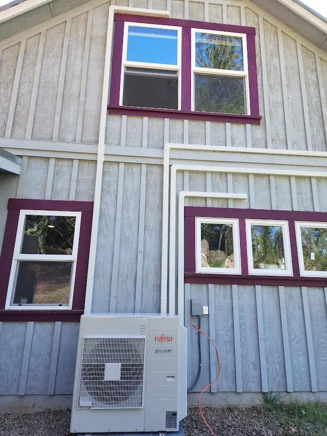 Gray wooden house exterior with maroon-trimmed windows; a white Fujitsu air conditioning unit is installed below.