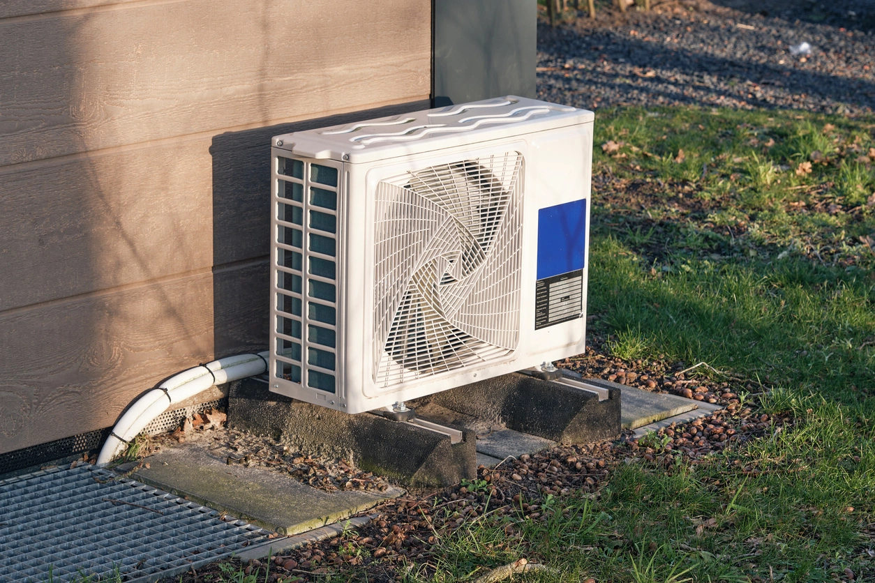 Outdoor heat pump unit next to a house, set on concrete slabs amidst grass and stones.
