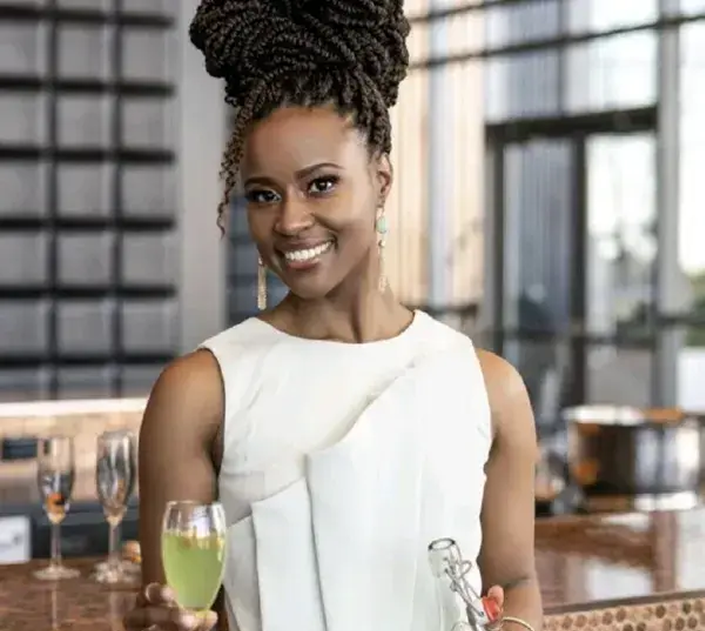 Smiling woman with braided hair styled up, wearing a white sleeveless dress, holding a glass of green drink in one hand and a bottle in the other, standing in a modern kitchen.