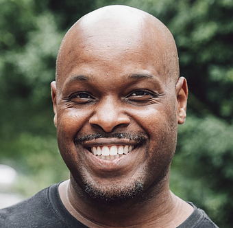 Smiling bald man with a goatee wearing a black shirt, with blurred green foliage in the background.