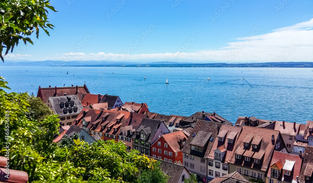 Blick auf traditionelle Dächer eines Städtchens am Ufer eines großen Sees mit Segelbooten und blauem Himmel.