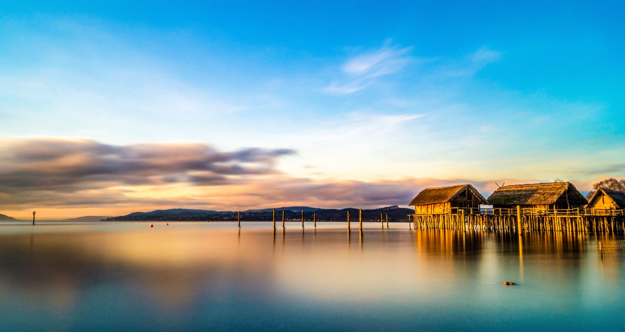Stelzenhäuser am ruhigen Seeufer bei Sonnenuntergang mit blauem Himmel und reflektierendem Wasser.