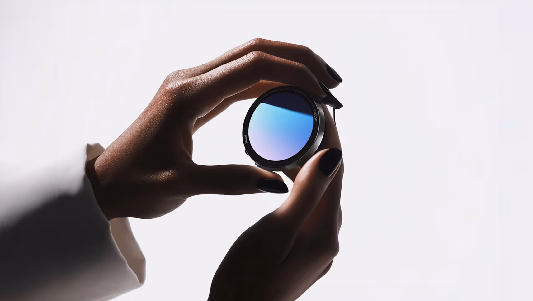 Two hands with black nail polish holding a round, reflective camera lens filter against a white background.