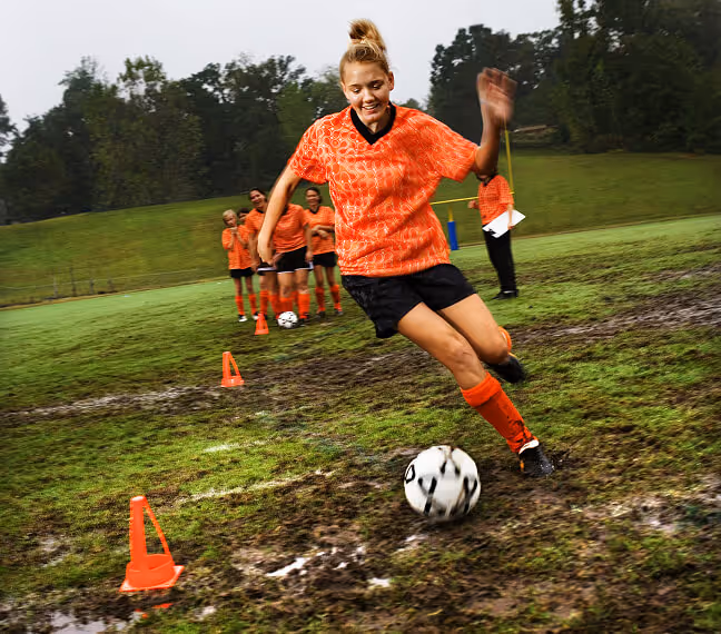 Ein Mädchen in orangenem Trikot dribbelt einen Fußball auf einem matschigen Spielfeld, während andere Kinder im Hintergrund zuschauen.