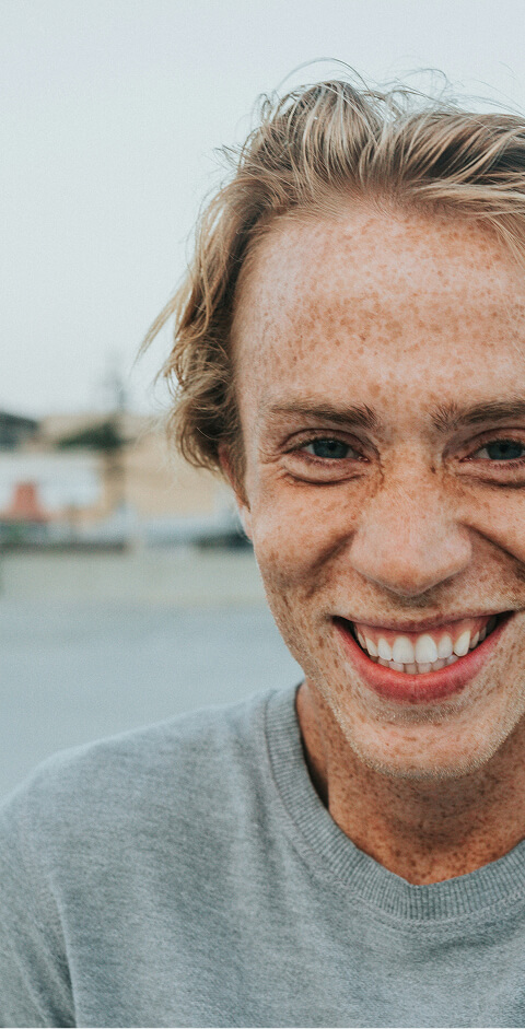 Bild Nahaufnahme eines lächelnden jungen Mannes mit Sommersprossen und blonden Haaren, der einen grauen Pullover trägt.