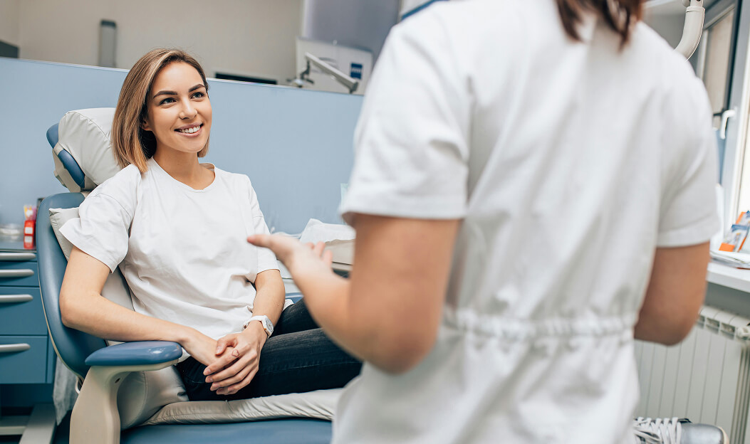 Bild Frau sitzt lächelnd auf einem Behandlungsstuhl im Gespräch mit einer medizinischen Fachkraft im Behandlungszimmer.