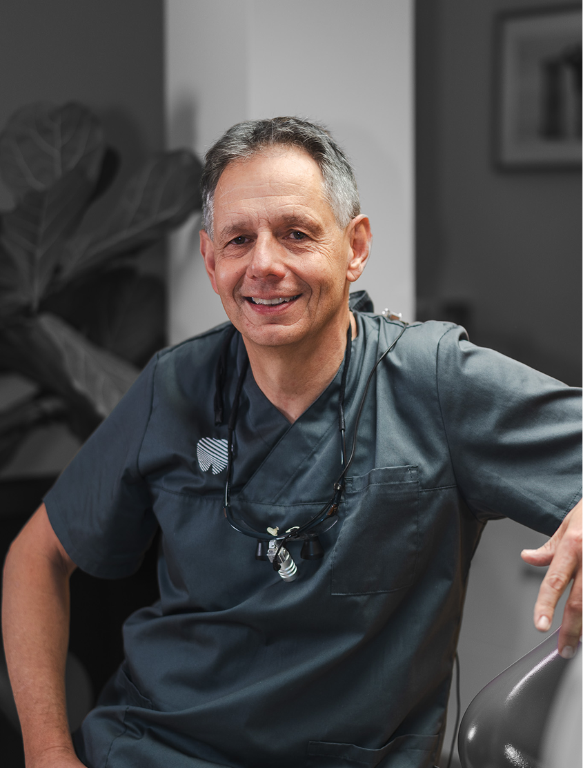 Bild Ein lächelnder Zahnarzt in grauer Arbeitskleidung sitzt in einer Praxis mit einer dentalen Lupe um den Hals.