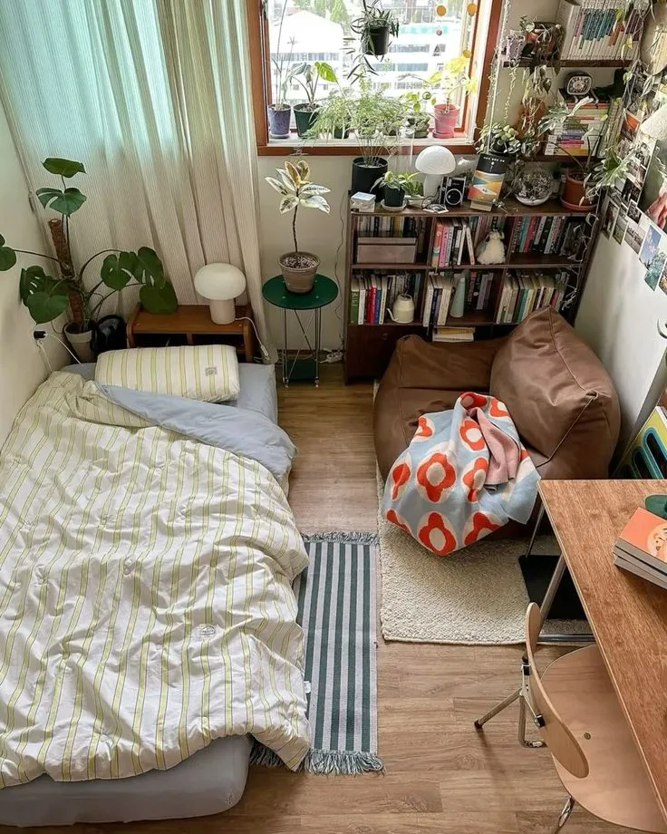 Cozy bedroom with a striped bed, brown armchair, plants, bookshelf, and wooden desk with chair.