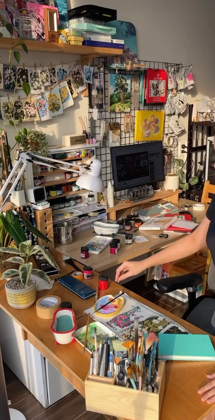 Art workspace with paints, brushes, botanical paintings, and a computer monitor on a wooden desk.