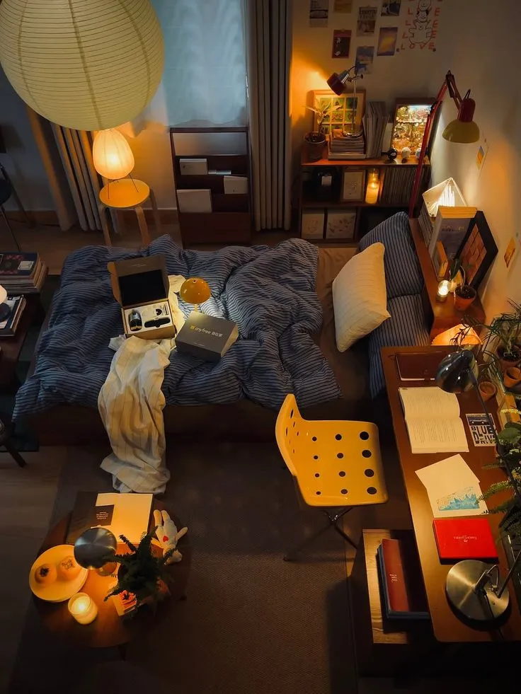 Cozy bedroom with blue striped bed, yellow chair, warm lamps, desk with books, and soft lighting.