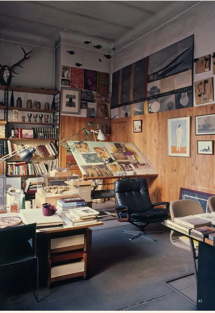 Cozy artist studio with wooden walls, drafting table, bookshelves, art prints, and black leather chair.