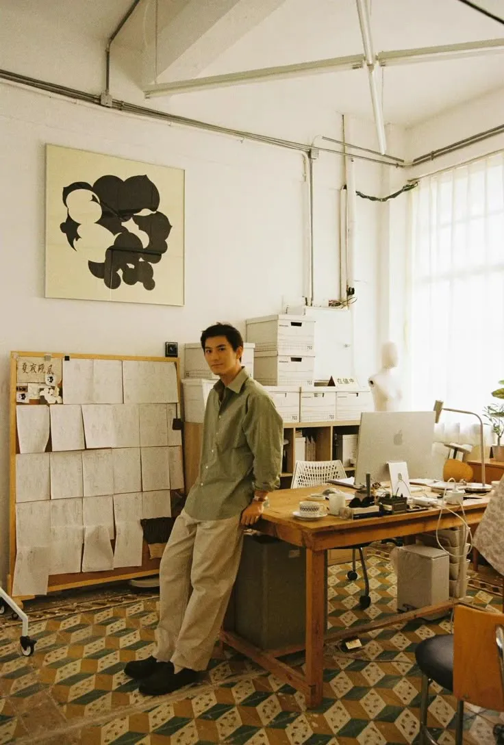 Man in green shirt and beige pants leans on a wooden desk in a sunlit office with papers and boxes.