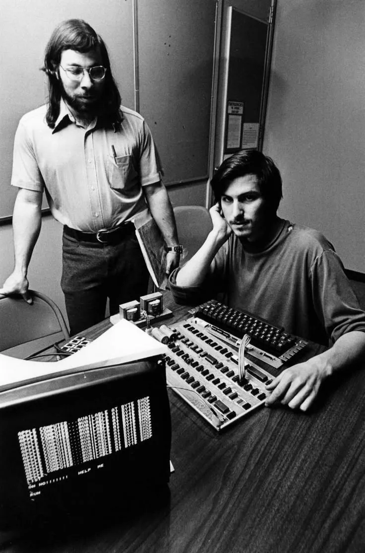Black and white photo of two men with early computer hardware and a monitor showing code.