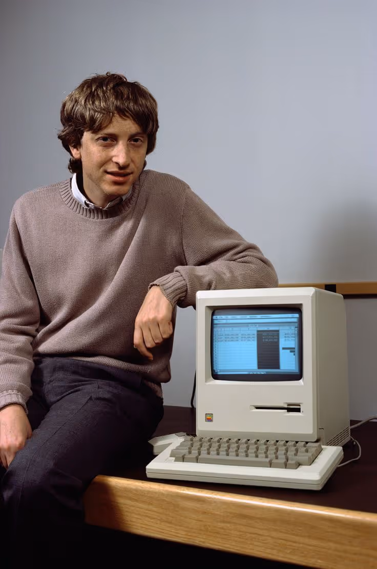 Young man in a brown sweater sitting on a desk beside a vintage Macintosh computer with spreadsheet on screen.