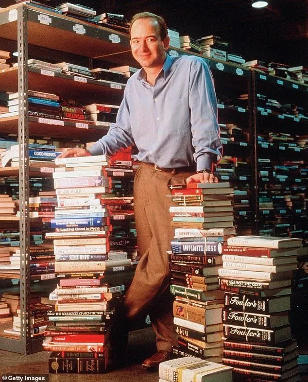 Man in blue shirt and brown pants stands smiling between two tall stacks of books in a library.
