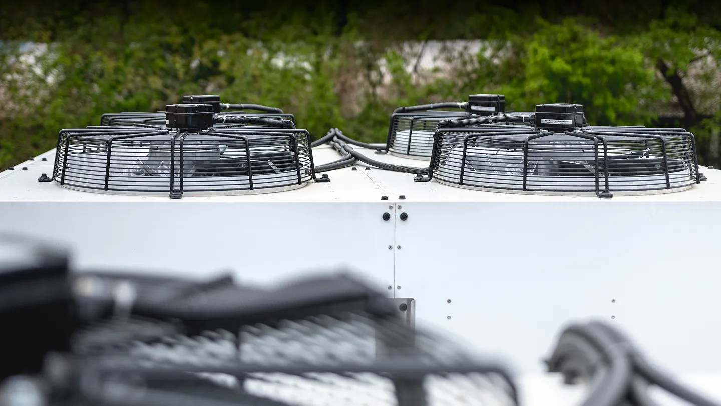 Průmyslový chladič s několika černými ochrannými mřížkami a ventilátory na bílém kovovém povrchu před zeleným pozadím stromů.