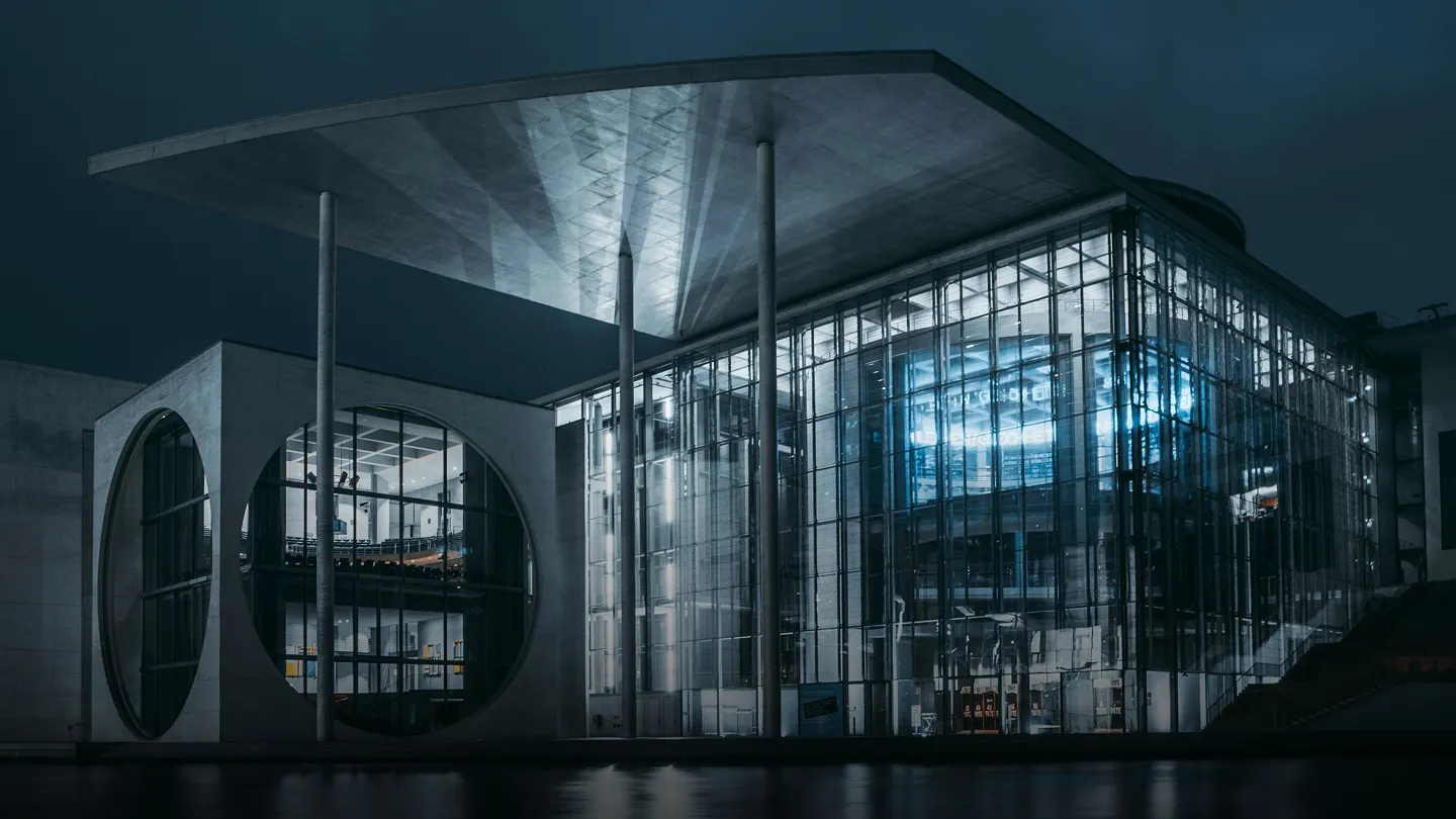 Moderní budova s velkými skleněnými stěnami a kruhovým oknem, osvětlená v noci.