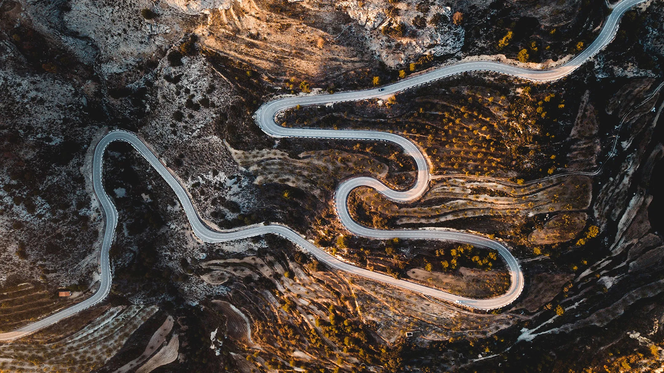 Záběr ze vzduchu na klikatou silnici vedoucí přes skalnatý a suchý terén s rozptýlenými keři a skalami.