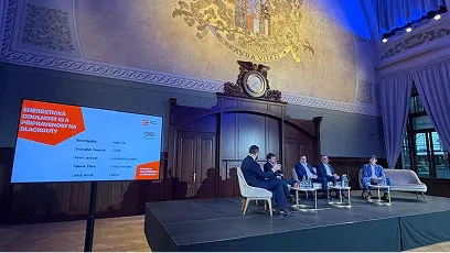 Čtyři muži sedí na pódiu v elegantní místnosti s dřevěným obložením a velkým hodinami, vedle nich je obrazovka s prezentací.