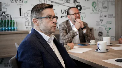 Muž ve tmavě modrém saku a brýlích sedí u stolu v zasedací místnosti s dalšími lidmi, kteří poslouchají.