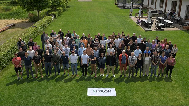 Skupina přibližně šedesáti lidí stojících na trávníku venku s praporkem ALTRON položeným před nimi.