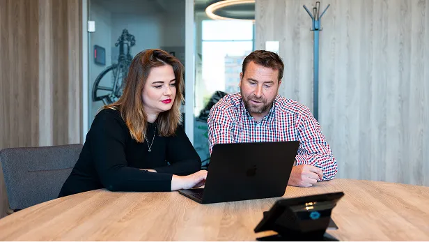 Dva lidé sedí u stolu a společně pracují na notebooku v moderní kanceláři.