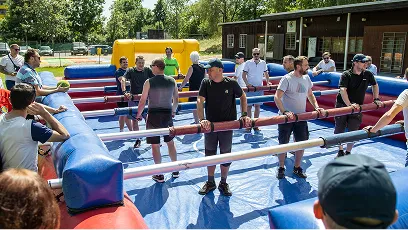 Skupina lidí hrajících obří stolní fotbal venku na nafukovacím hřišti za slunečného dne.