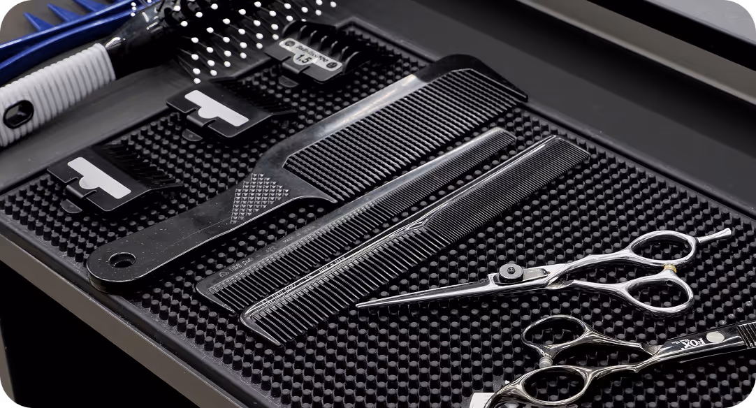 Hairdressing tools including two metal scissors, two black combs, and a hairbrush with a white handle on a textured black mat.