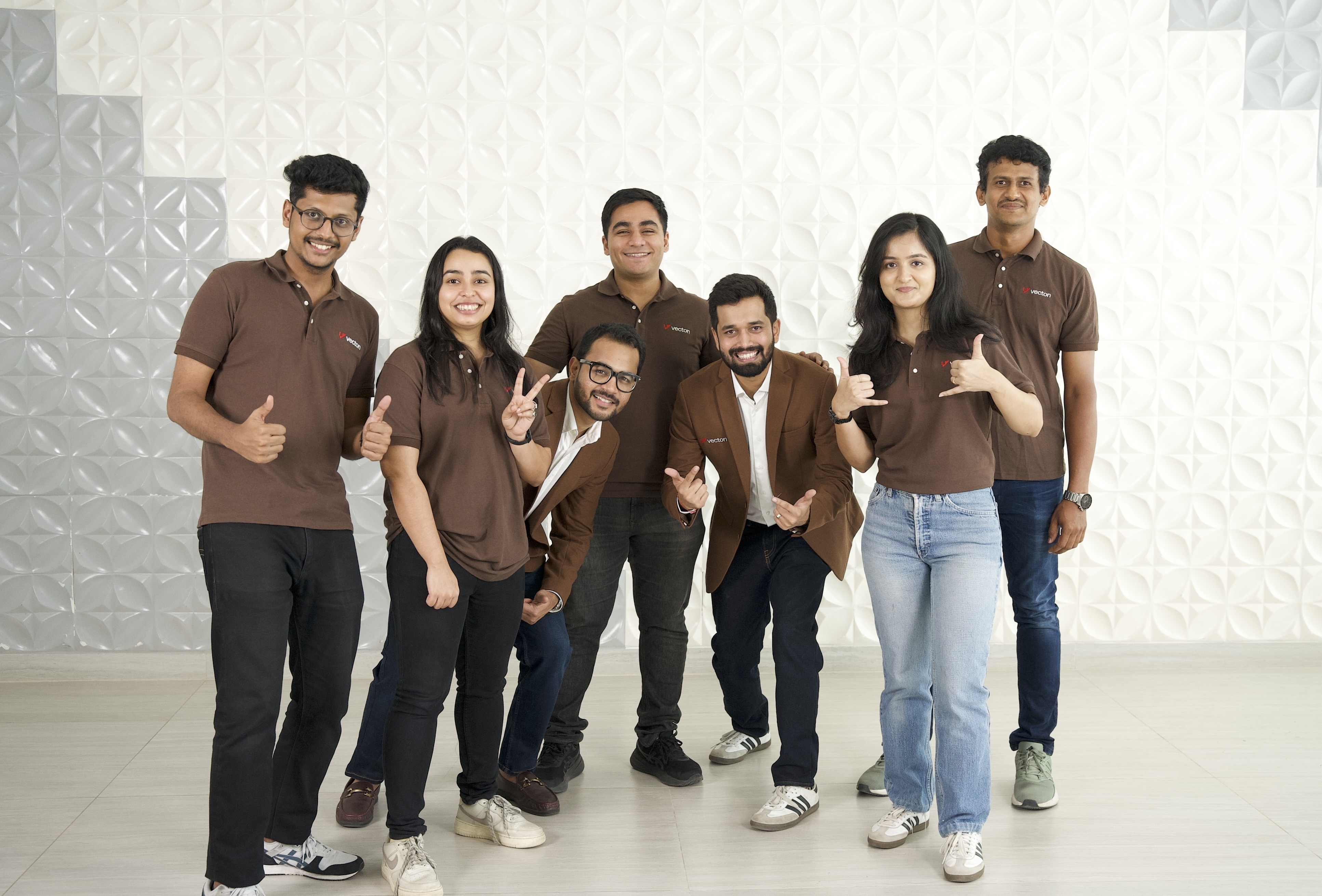 Seven people smiling and posing with hand gestures against a textured white wall background.