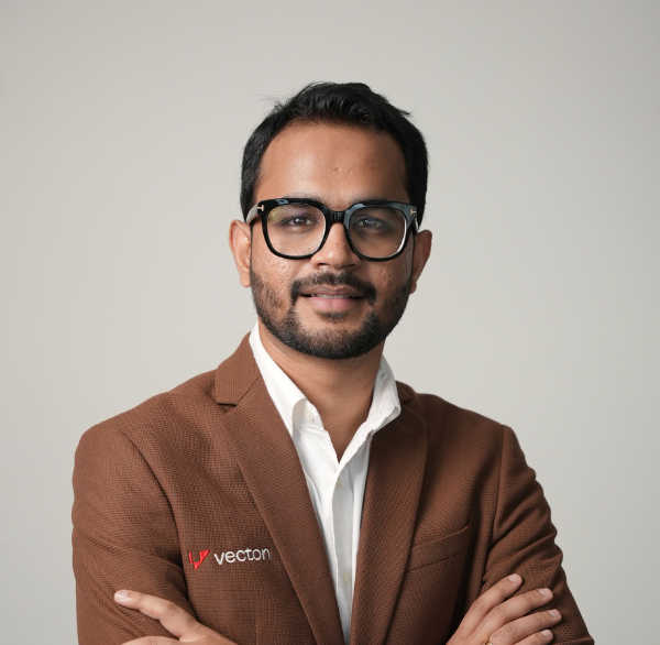 Man with glasses and beard wearing a brown blazer and white shirt, arms crossed, smiling slightly.
