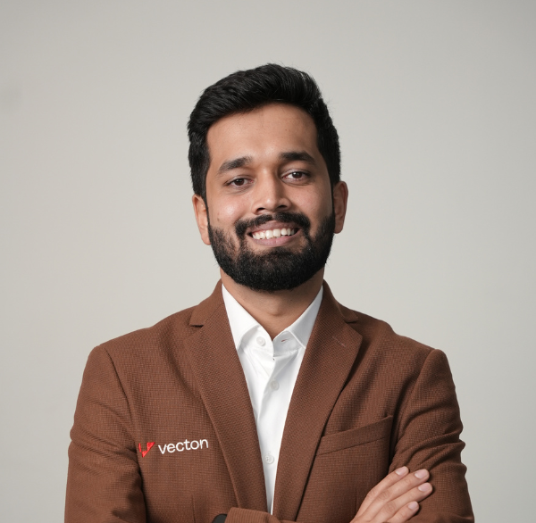 Smiling man with beard wearing a brown blazer with 'Vecton' logo and white shirt on gray background.