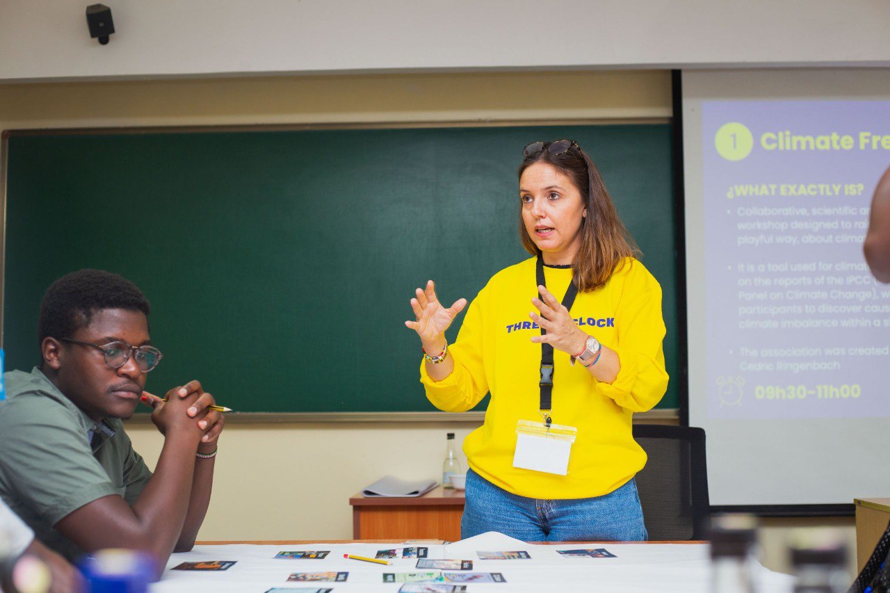 Woman in yellow sweatshirt explaining with hand gestures in a classroom while a man listens attentively.