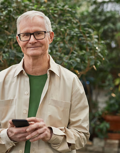 Smiling elderly man with glasses wearing a beige shirt and green t-shirt holding a smartphone outdoors with green foliage in the background.