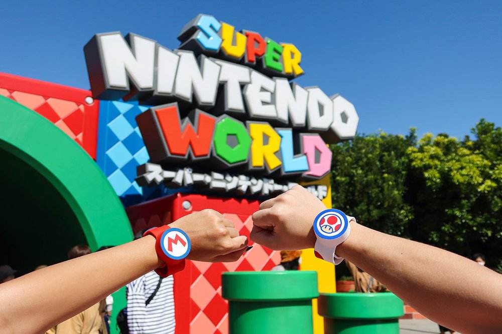 Două mâini în prim-plan poartă brățări cu simboluri Mario și Toad în fața semnului colorat Super Nintendo World, cu țevi verzi și copaci în fundal.