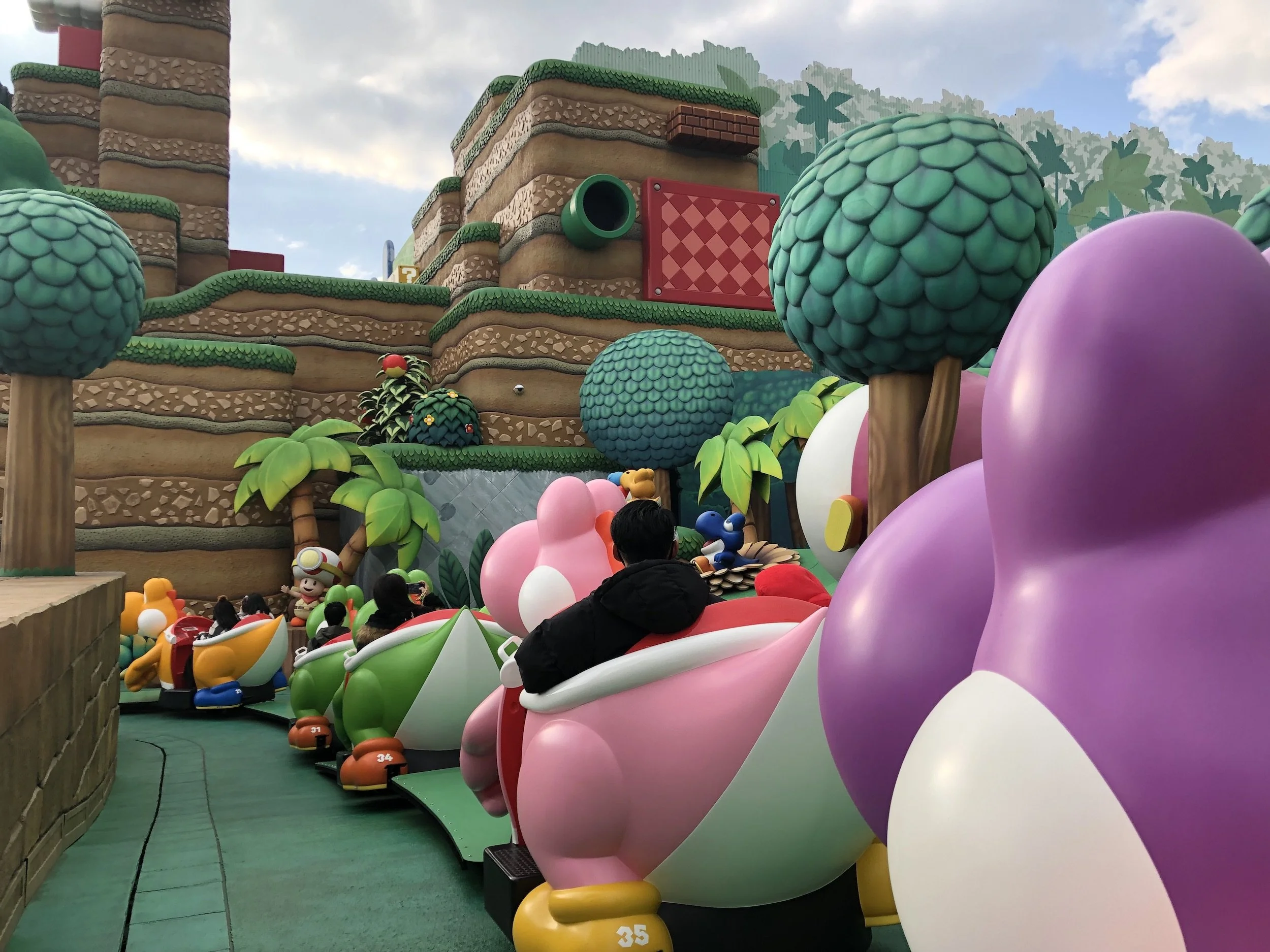 Persoană într-un carusel cu tematică Yoshi, cu vehicule colorate și un decor cu copaci și munți stilizați la Super Nintendo World.